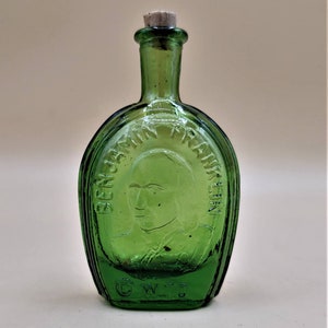 May include: A green glass bottle with a cork stopper. The bottle is shaped like a horseshoe and has a raised image of Benjamin Franklin on the front. The bottle is marked with the letters "C.W.T."