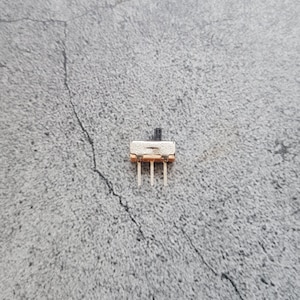 May include: A small, white, rectangular toggle switch with a black lever. The switch has three pins on the bottom.