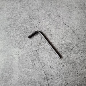 Op de afbeelding: Een zwarte metalen inbussleutel, ook wel een zeskantsleutel genoemd, ligt op een grijs, getextureerd oppervlak.