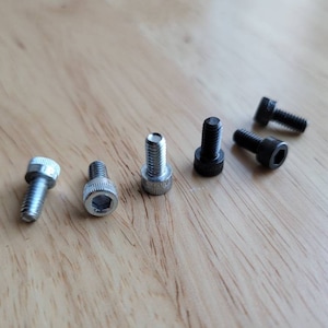 May include: A close-up of six screws, five silver and one black, on a light wood surface.