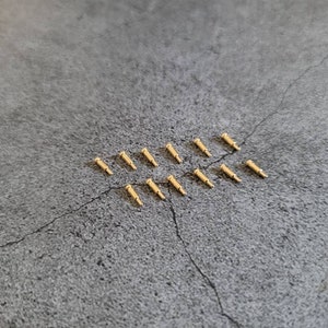 May include: A close-up of a group of 12 gold-colored metal pins, arranged in a row on a gray textured surface.