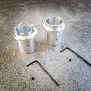 May include: Two silver metal parts with a hexagonal shape and a small hole in the center. The parts are on a gray surface with two black Allen wrenches.