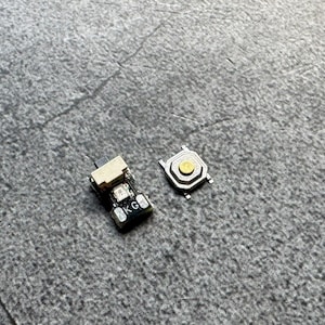 Pode incluir: Dois pequenos componentes eletrônicos, um botão preto e bege com as letras "BKG" e um botão prateado e dourado, são exibidos em uma superfície cinza texturizada. Estes componentes são provavelmente usados em eletrônica.