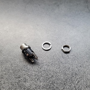 May include: A close-up of a black electrical connector with two silver metal rings. The connector is on a gray surface.