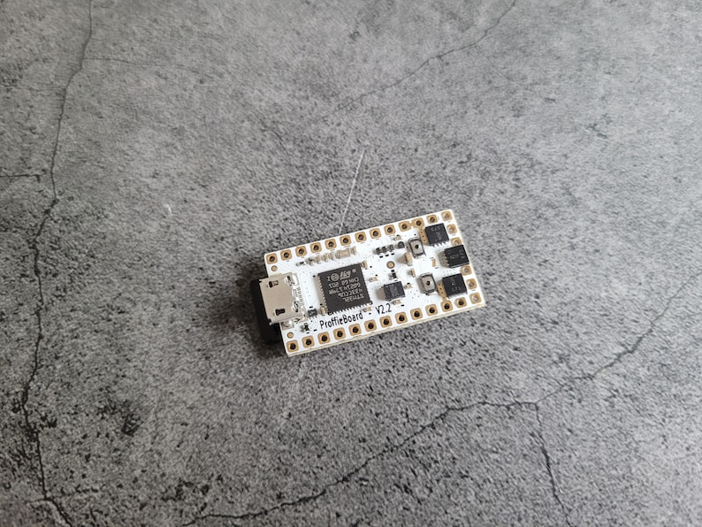 Puede incluir: Una placa de circuito blanco con pines de color dorado y un conector USB negro. La placa tiene un chip negro con el texto "ProffieBoard V2.2" impreso en &eacute;l.