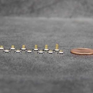 May include: A close-up of a row of small, silver-colored, electronic switches with gold-colored contacts. The switches are arranged in a line on a gray surface. A copper-colored coin is in the background.