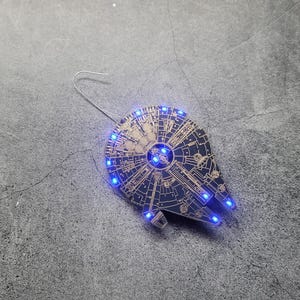 May include: A black and gold ornament shaped like the Millennium Falcon spaceship from Star Wars. The ornament has blue LED lights around the edges.