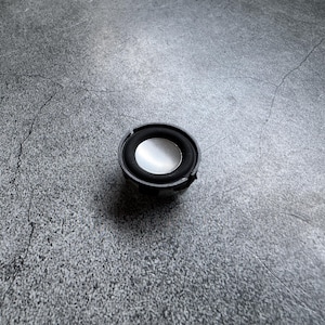 May include: A small, round speaker with a black frame and a silver center. The speaker is positioned on a gray, textured surface. The speaker is a component of electronics.