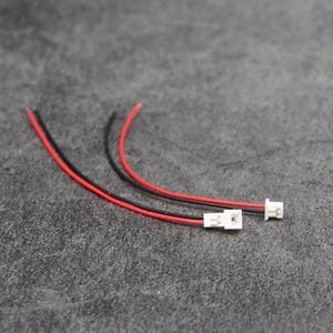 Könnte beinhalten: Zwei weiße elektrische Stecker mit roten und schwarzen Drähten. Die Stecker befinden sich auf einer grauen Oberfläche.