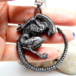 Op de afbeelding: Een zilverkleurige hangerketting met een gedetailleerd Xenomorph-ontwerp. De hanger toont het wezen in een opgerolde pose, met ingewikkelde skelet- en getextureerde details. De ketting heeft een delicate ketting en een veilige sluiting.