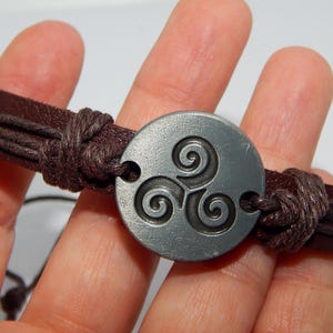 May include: A brown leather bracelet with a circular, silver-colored centerpiece. The centerpiece features a Celtic triskele design. The bracelet is held together with knotted leather cords.