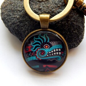 May include: A bronze key chain with a round glass charm featuring a colorful Aztec-inspired design. The design is a stylized face with blue, red, black, and white details.