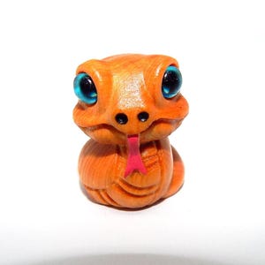 May include: A carved wooden snake figurine with an orange body, black nostrils, and a pink tongue. The snake has large, bright blue eyes. The figurine is a whimsical representation of a snake.