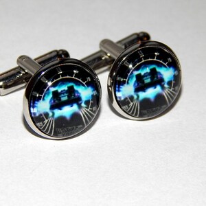 Puede incluir: Un par de gemelos de plata con una imagen en negro y azul de un velocímetro de coche con la aguja a 88 mph. La imagen está rodeada por un anillo de plata.