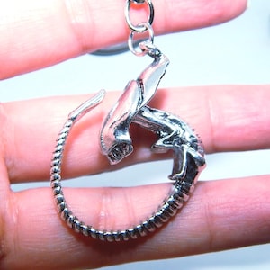 May include: A silver-colored pendant in the shape of an alien facehugger. The pendant is attached to a silver chain. The facehugger has a detailed design, with a textured body and a menacing expression. The pendant is a unique piece of jewelry.