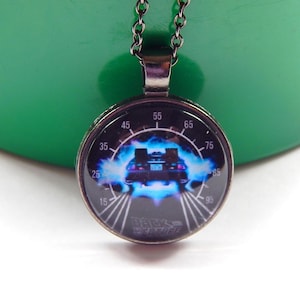 Puede incluir: Un colgante redondo con marco de metal negro y cabujón de cristal. El gráfico muestra un coche rodeado de luz azul sobre un dial de velocímetro con números del 15 al 95 y el texto Back to the Future en la parte inferior del colgante.