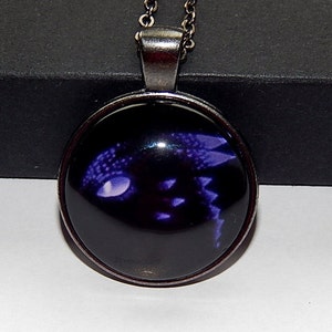 May include: A silver necklace with a round glass pendant featuring a purple dragon silhouette on a black background.