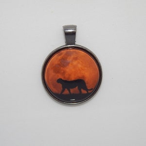 K&ouml;nnte beinhalten: Ein silberner Anh&auml;nger mit einem runden Glascabochon, der eine Silhouette eines Geparden zeigt, der vor einem rot-orangen Mond geht.