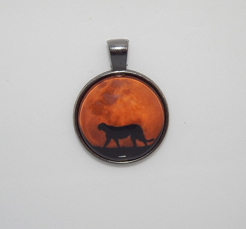 K&ouml;nnte beinhalten: Ein silberner Anh&auml;nger mit einem runden Glascabochon, der eine Silhouette eines Geparden zeigt, der vor einem rot-orangen Mond geht.