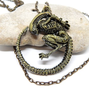 May include: A bronze-colored pendant necklace featuring a detailed Alien design. The pendant depicts the creature in a circular arrangement, with a chain attached for wearing. The necklace is set against a neutral stone background.