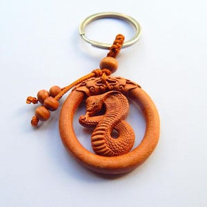Puede incluir: Un llavero de madera con un diseño de cobra tallada. La cobra está dentro de un marco circular, con escamas detalladas y una capucha levantada. El llavero incluye un anillo de metal, un cordón decorativo y cuentas de madera. La madera tiene un tono marrón rojizo cálido.