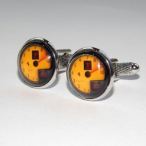 Può includere: Un paio di gemelli d'argento con un design circolare giallo e nero. Il design presenta un tachimetro con i numeri da 1 a 10 e le lettere S e 300.