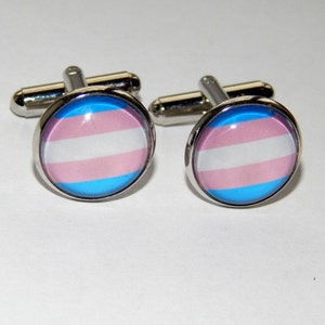 Könnte beinhalten: Ein Paar silberne Manschettenknöpfe mit einem runden, blauen, rosa und weißen Design. Das Design ist eine Darstellung der Transgender-Pride-Flagge.