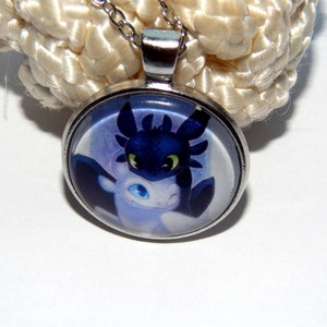 May include: A silver necklace with a round glass pendant featuring a cartoon image of a black and white dragon. The dragon has green eyes and is set against a purple and blue background.
