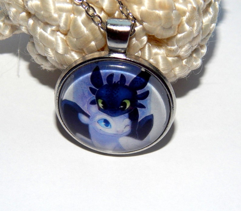 May include: A silver necklace with a round glass pendant featuring a cartoon image of a black and white dragon. The dragon has green eyes and is set against a purple and blue background.