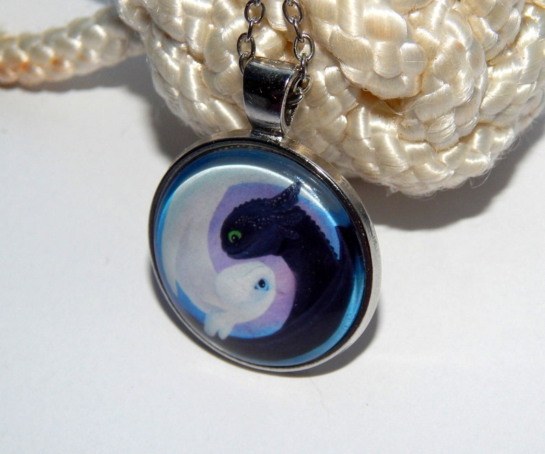 May include: A silver necklace with a round glass pendant featuring a black and white dragon design. The dragons are in a yin yang symbol with a blue background.