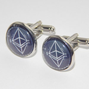 May include: A pair of silver cufflinks with a blue and white Ethereum cryptocurrency symbol. The cufflinks have a round face with a shiny silver border and a T-bar closure.