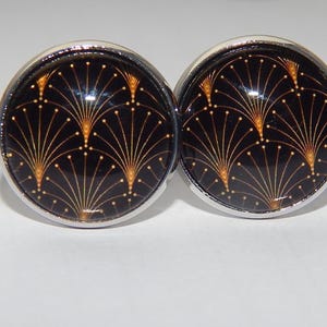 Puede incluir: Un par de gemelos redondos con fondo negro y diseño de abanico art déco dorado. Los gemelos tienen un borde de metal plateado y un acabado brillante. Accesorios adecuados para ropa formal.
