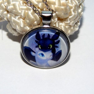 May include: A silver necklace with a round glass pendant featuring a cartoon image of a black and white dragon. The dragon has green eyes and is depicted in a close-up view.