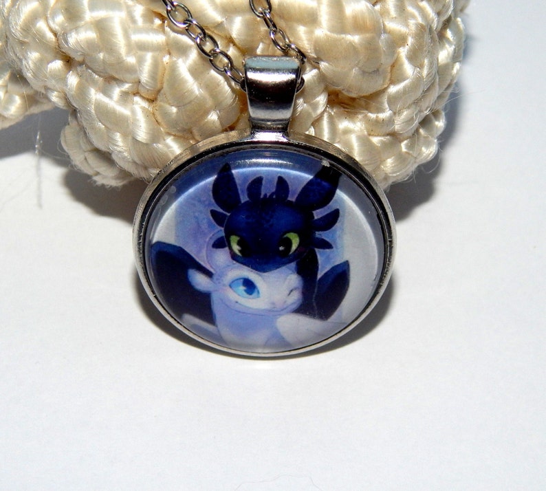 May include: A silver necklace with a round glass pendant featuring a cartoon image of a black and white dragon. The dragon has green eyes and is depicted in a close-up view.