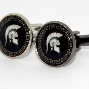Könnte beinhalten: Ein Paar Manschettenknöpfe mit schwarzem Hintergrund und einem weißen Bild eines spartanischen Helms mit einem griechischen Schlüsselrand. Ein Manschettenknopf ist silberfarben und der andere ist schwarz.