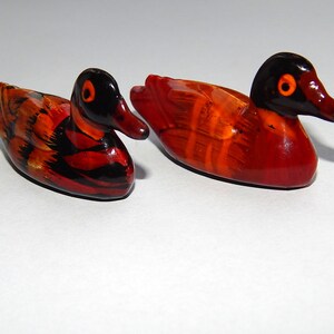 Puede incluir: Dos figuras decorativas de patos. Los patos tienen cabezas negras con ojos y picos naranjas. Los cuerpos están pintados en tonos de rojo, naranja y negro. Las figuras son pequeñas y parecen estar hechas de un material brillante.