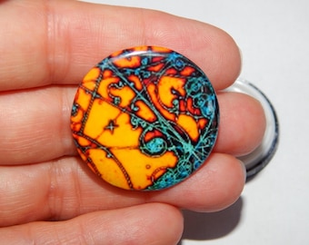 May include: A round button with a colorful abstract design. The design features a yellow background with blue, red, and black swirls and lines.