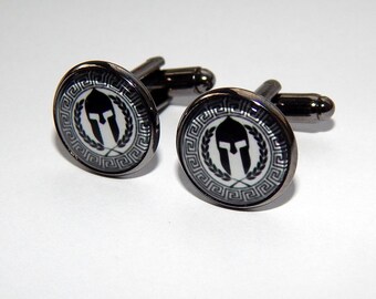 May include: A pair of black cufflinks with a white and black image of a Spartan helmet with a laurel wreath and a Greek key border.