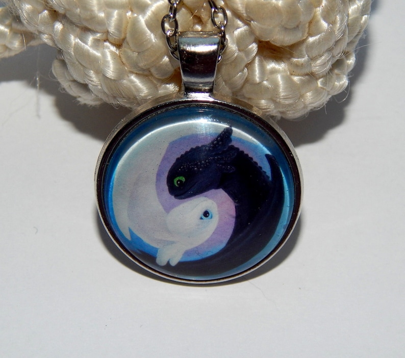 May include: A silver necklace with a round glass pendant featuring a black and white dragon design. The dragons are in a yin yang symbol with a blue background.