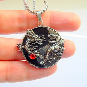 Puede incluir: Collar con colgante de plata con un diseño de yin yang con un dragón y un tigre sobre un fondo negro. Una piedra preciosa roja está en el centro del colgante.