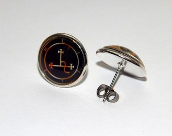Könnte beinhalten: Ein Paar silberne Ohrstecker mit einem runden Design in Schwarz und Orange. Das Design zeigt ein Symbol mit einem Kreuz und einer Schlange.