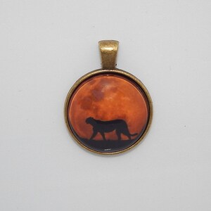 K&ouml;nnte beinhalten: Ein Bronzependant mit einem runden Glas-Cameo, das eine Silhouette eines Geparden zeigt, der &uuml;ber einen rot-orangen Mond l&auml;uft.