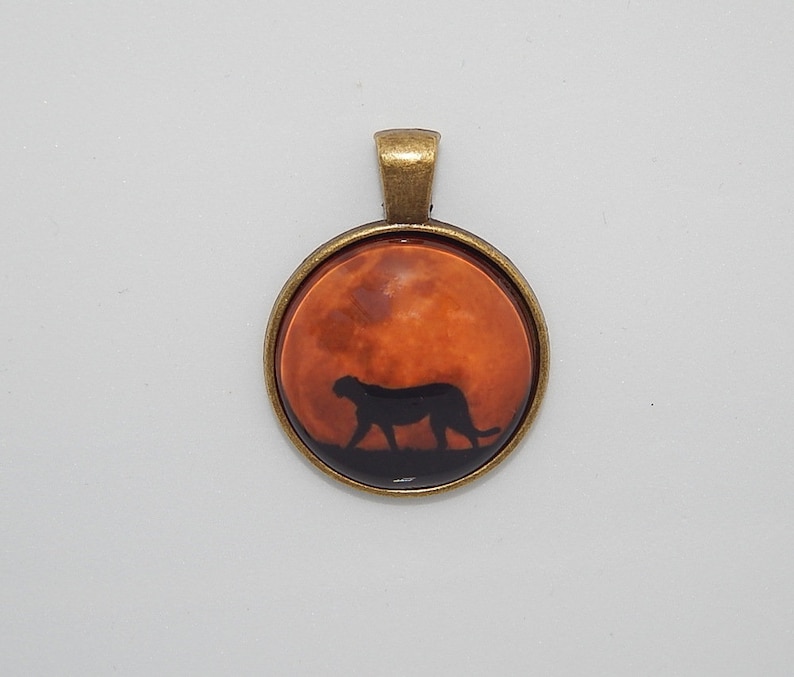 K&ouml;nnte beinhalten: Ein Bronzependant mit einem runden Glas-Cameo, das eine Silhouette eines Geparden zeigt, der &uuml;ber einen rot-orangen Mond l&auml;uft.