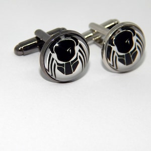 May include: A pair of silver cufflinks with black and white enamel designs. The design features a stylized creature with a black head and white wings.