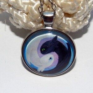 May include: A silver necklace with a round pendant featuring a yin yang design with a black and white dragon on each side. The dragons are facing each other and have green eyes.