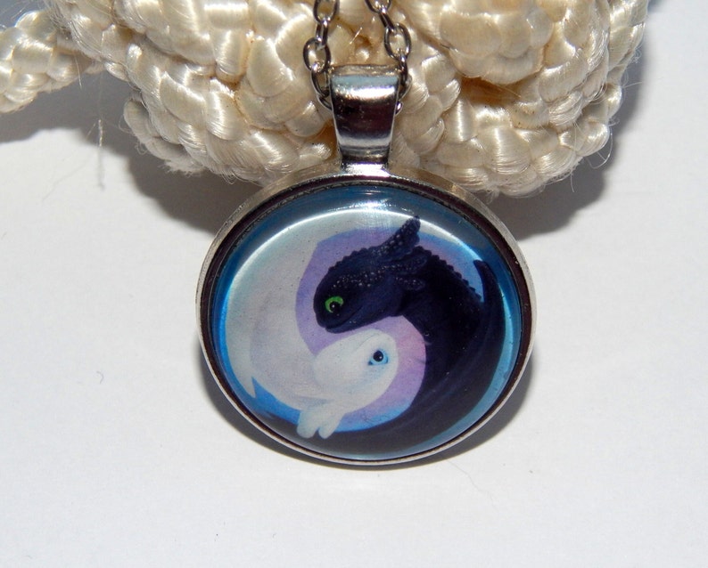May include: A silver necklace with a round pendant featuring a yin yang design with a black and white dragon on each side. The dragons are facing each other and have green eyes.