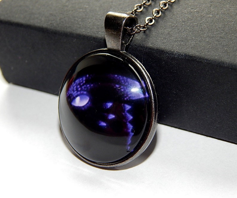 May include: A black necklace with a round glass pendant featuring a purple dragon silhouette. The pendant has a silver metal frame.