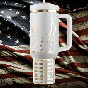 以下が含まれることがあります： ハンドル、ゴールドのアクセント、蓋が付いた白い断熱タンブラー。タンブラーには、ドッグタグと「Officer」の文字がデザインされています。背景はアメリカ国旗です。