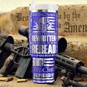 May include: A blue and silver travel mug with the text "We the People" and "It doesn't need to be rewritten, it needs to be reread." The mug features a skull and crossbones design and American flag imagery. Firearms and ammunition are in the background.