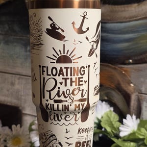 May include: Cream-colored travel mug with a bronze-colored rim and lid. The mug features brown graphics of kayaks, anchors, and the text "Floating the River & Killin' My Liver." Additional text includes "Drinking at the River" and "Keeping it Reel at the River."